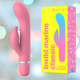 A pink dual motor vibrator, the B Swish Bwild Classic Marine Rabbit Vibrator in Guava, is displayed next to its colorful box adorned with the text "B Swish" and "Bwild Classic Marine." The packaging features a gradient design with illustrations against a bright yellow background. The scene is set with a blurred, colorful bokeh effect in the backdrop.